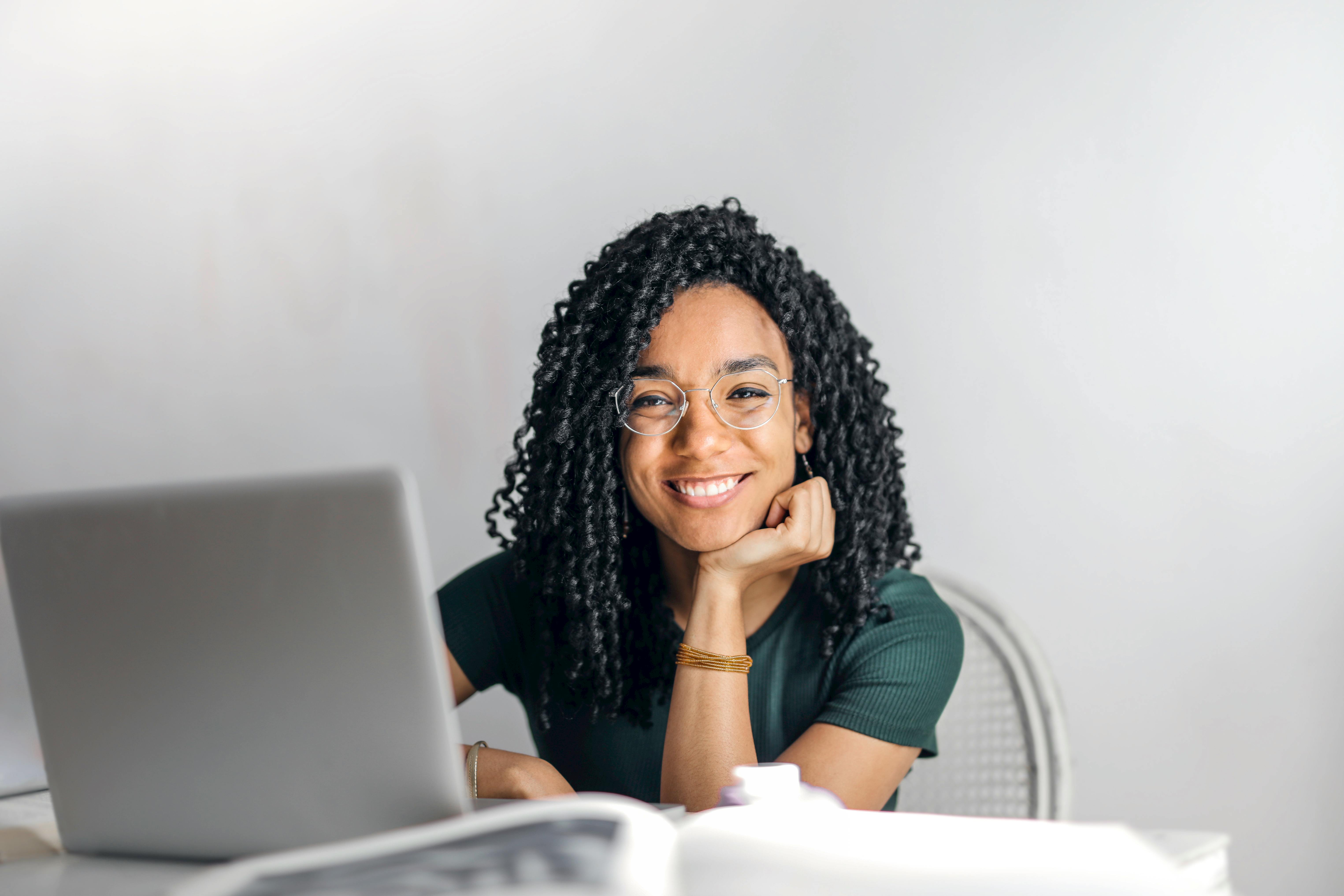 a black woman with long curly black hair and glasses and dark green tshirt smiles at the camera from a laptop