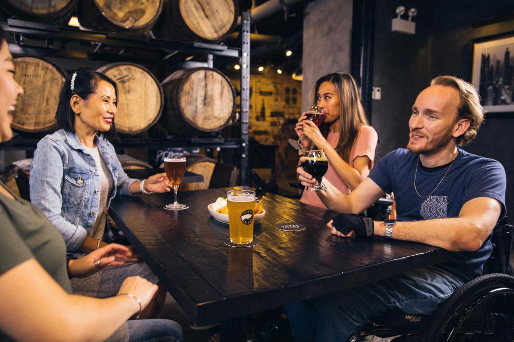 a group of 4 young adults enjoying a beer at a table at a brewery. One of the individuals is a wheelchair user.
